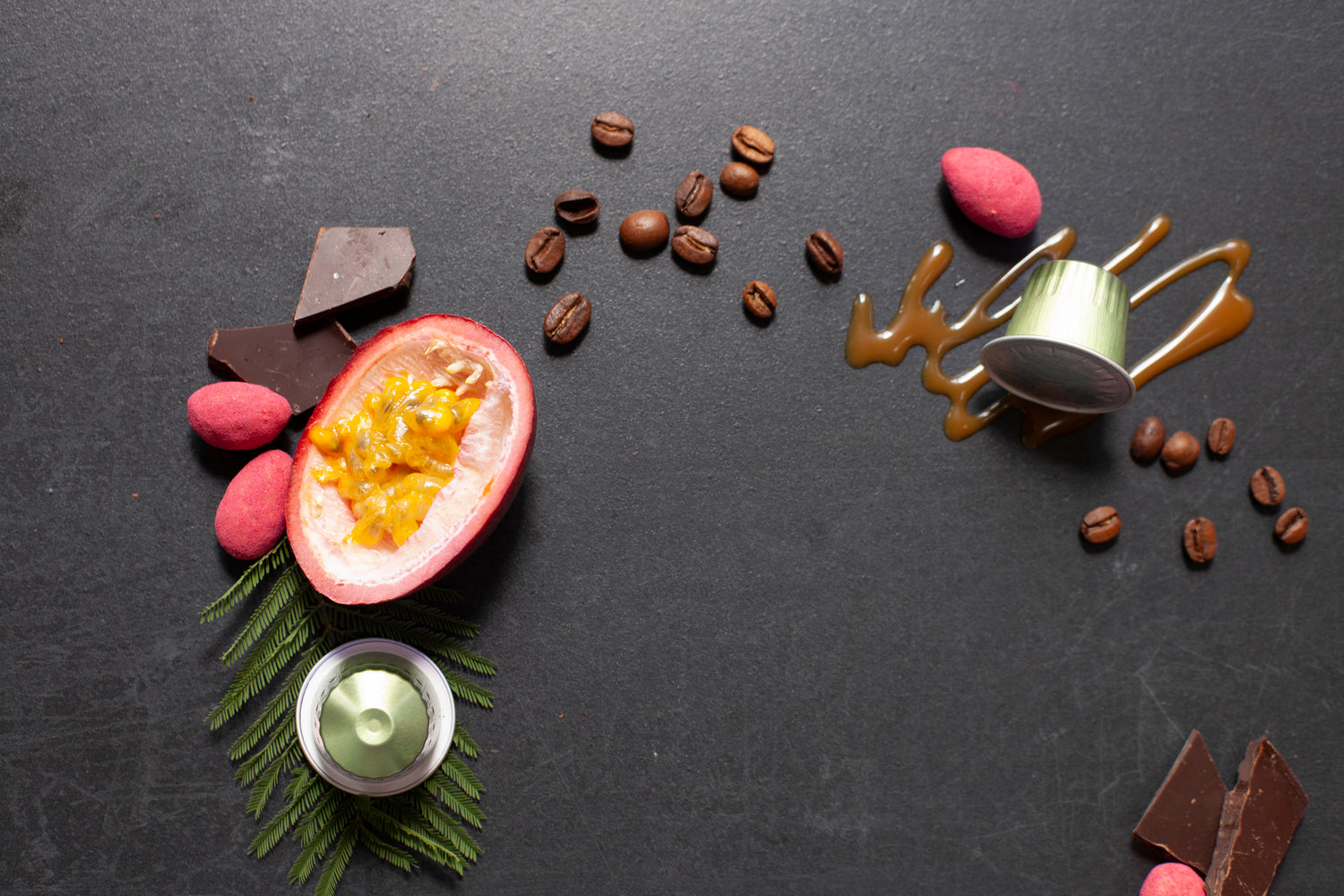Decorative arrangement of coffee beans, coffee capsules, chocolate pieces, and a halved passion fruit on a dark surface.
